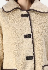 Beige schapenvachtjas met een ronde kraag, donkerbruine afwerking en vier zwarte knopen. Zachte, gestructureerde stof met een gezellige uitstraling.