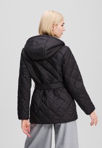 Veste matelassée noire avec capuche, ceinture à la taille et motif en losanges. Le tissu semble brillant et léger, idéal pour les temps frais.