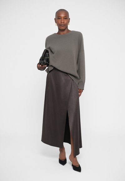 Mujer con un suéter gris holgado, falda envolvente marrón oscuro con abertura frontal, tacones negros de punta y sosteniendo un bolso negro de cadena.
