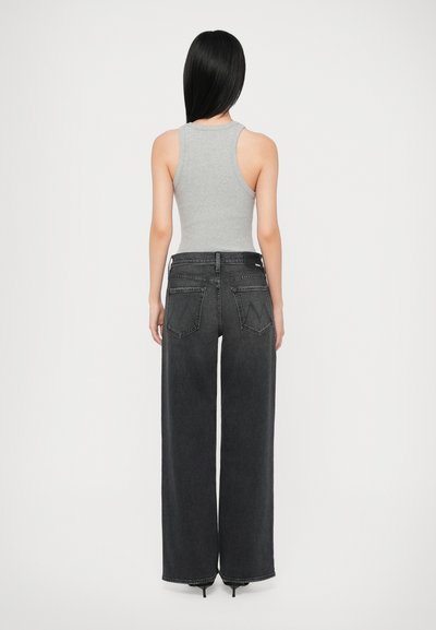 Ženska z dolgimi črnimi lasmi, oblečena v sivo brezrokavno majico, široke črne kavbojke in črne pete, stoji z obraza obrnjena stran od belega ozadja.