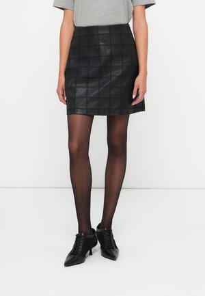 Femme portant une mini-jupe en cuir noire à carreaux, des collants noirs transparents, des chaussures à talons lacées noires et un haut gris à manches courtes.