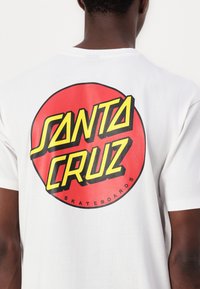Λευκό βαμβακερό T-shirt με μεγάλης διαστάσεως κόκκινο κυκλικό γραφικό που αναγράφει "SANTA CRUZ" με έντονα κίτρινα και μαύρα γράμματα, με την ένδειξη "SKATEBOARDS" από κάτω.