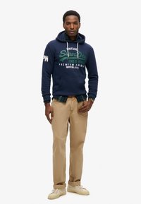 Felpa navy con logo verde e bianco, pantaloni beige e sneaker color crema. Strato di una camicia a scacchi sotto la felpa.
