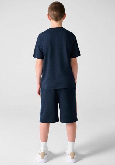 Niño de pie de espaldas, vistiendo una camiseta azul marino y pantalones cortos con rayas laterales de colores, calcetines blancos y zapatillas blancas.