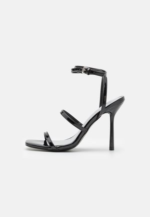 Sort patentlæder sandal med høje hæle og tynde stropper over tæerne, mellemfoden og spænde ved anklen, vist på hvid baggrund.