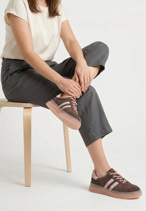 Persoon zittend op een houten krukje, draagt bruine en roze sneakers, grijze broek en een wit shirt met korte mouwen, past met beide handen een schoen aan.