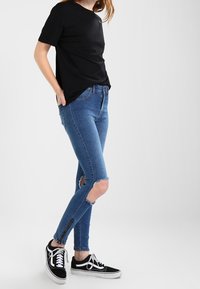 Black short-sleeve shirt paired with blue skinny jeans featuring knee rips and black sneakers with white laces, set against a light background.