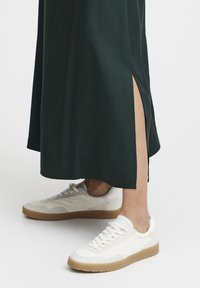Jupe verte, longueur genou, avec des fentes latérales, associée à des baskets blanches avec une semelle texturée beige et des accents subtils.