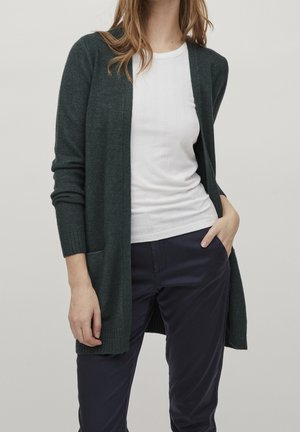 Cardigan verde scuro in morbido tessuto, con maniche lunghe, colletto a scialle e tasche frontali, indossato sopra un top bianco a coste.