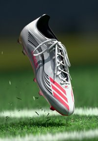Cizmă de fotbal albă cu accente roz și negre, șireturi și un design aerodinamic cu o suprafață texturată, ridicată deasupra ierbii verzi.