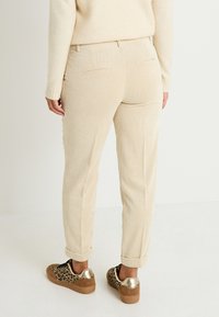 Pantalones de tergal beige con rayas verticales, con dobladillos acampanados y dos bolsillos traseros, combinados con zapatillas de leopardo y acentos dorados.