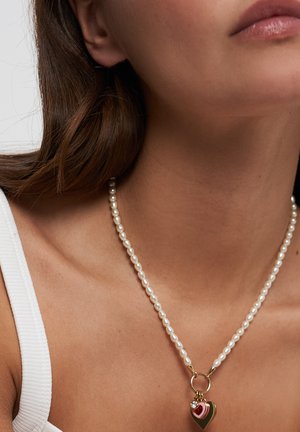 Vrouw draagt een wit hemdje en een parelketting met gouden ringen en gelaagde hartvormige hangers in rood, roze en goud.