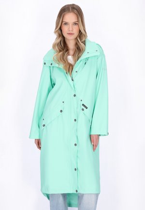 Fiatal nő hosszú, hullámos szőke hajjal, mentazöld hosszú esőkabátot visel, gombos és cipzáras záródással, világoskék farmernadrág fölött.