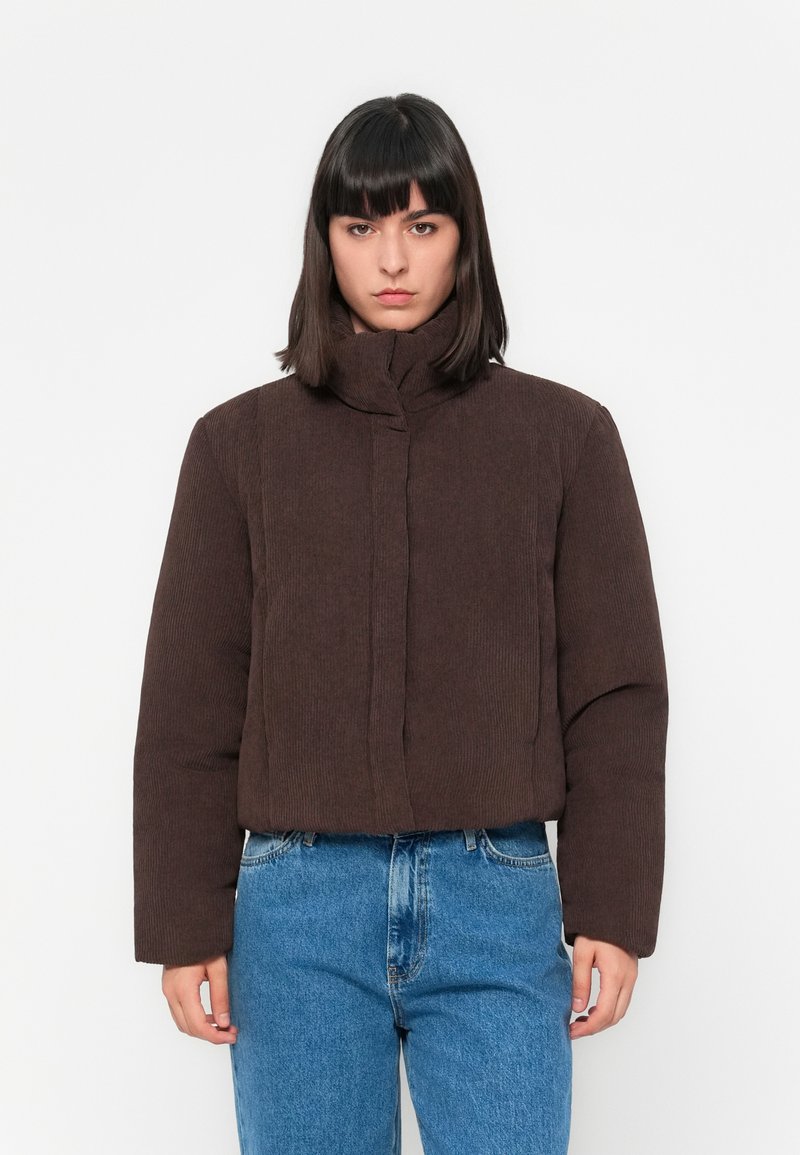 Donna con capelli scuri di lunghezza spalle, che indossa un giubbotto imbottito marrone e jeans blu, in piedi davanti a uno sfondo bianco semplice.