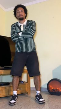Zelena črtasta rugby majica z dolgimi rokavi, črne jeans kratke hlače, črne teniske z belimi detajli, stoji s križanimi rokami v dnevni sobi.