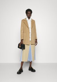 Beige wollen jas met gesneden revers, witte blouse met kleurrijke knopen, beige en blauw gestreepte rok, zwarte loafers en zwarte handtas.