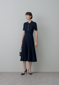Mornarsko modra rebrasta obleka z ovratnikom, kratkimi rokavi, zlatimi gumbi in plisiranim krilom. Kombinirana s temno modrimi petkami in majhno črno torbico.