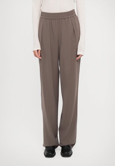 Persona vistiendo pantalones de pierna ancha en color topo con cintura fruncida, blusa blanca de manga larga y zapatos negros de punta cuadrada, de pie sobre un fondo blanco.