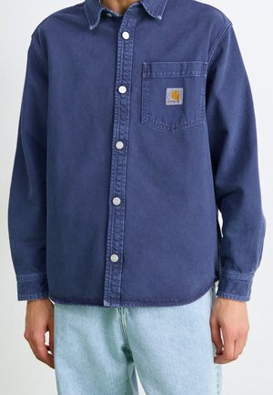 Personne portant une chemise en jean bleu foncé boutonnée avec un logo sur la poche poitrine et un jean bleu clair, debout les bras détendus.