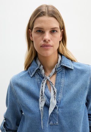 Blondynka w jasnej dżinsowej koszuli z szalikiem w szary kwiatowy wzór wiązanym wokół szyi, patrząca przed siebie z neutralnym wyrazem twarzy.