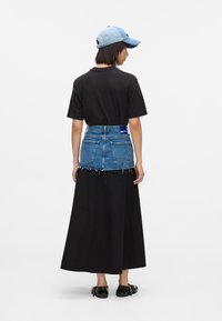 Fekete pólót viselő személy, amely a hosszú fekete szoknyába van tűrve, rövid farmerelővel, kék baseball sapkával és fekete cipővel, háttal áll.