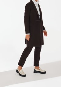 Manteau long marron avec deux boutons, associé à un pantalon foncé et des mocassins crème. Présente un design élégant, des textures contrastantes et des semelles épaisses.