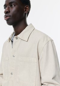 Beige denim skjortejakke med to forlommer, krave og sølvfarvede trykknapper. Stoffet ser tekstureret og let holdbart ud.