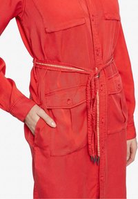 Robe-chemise rouge avec poches à rabat sur le devant et ceinture assortie nouée à la taille, main posée dans une poche.