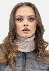 Prim-plan al unei persoane îmbrăcate cu un pulover cu guler înalt, de culoare roz deschis, sub o geacă groasă și matlasată, de culoare închisă. Părul este ondulat și brunet.