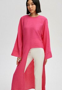 Túnica larga y holgada de tejido texturizado en un vivo color rosa, con mangas anchas y una gran abertura en un lado. Combinada con pantalones blancos.