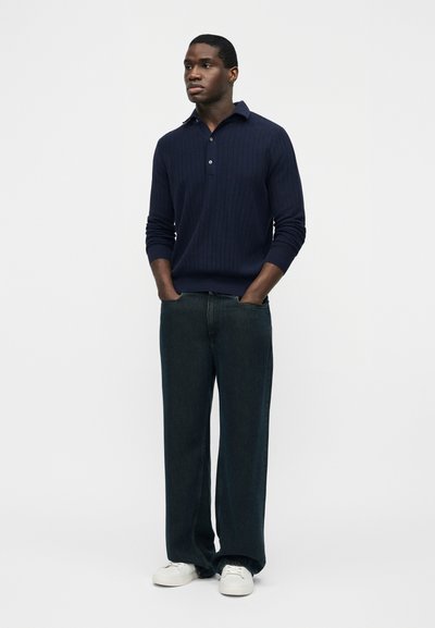 Homme debout, les mains dans les poches, portant un polo manches longues bleu marine, un jean large foncé et des baskets blanches, sur fond blanc.