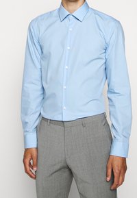 Lichtblauw overhemd met lange mouwen, voorzien van een puntige kraag en witte knopen. Draagt een grijze dresscode broek met textuur.