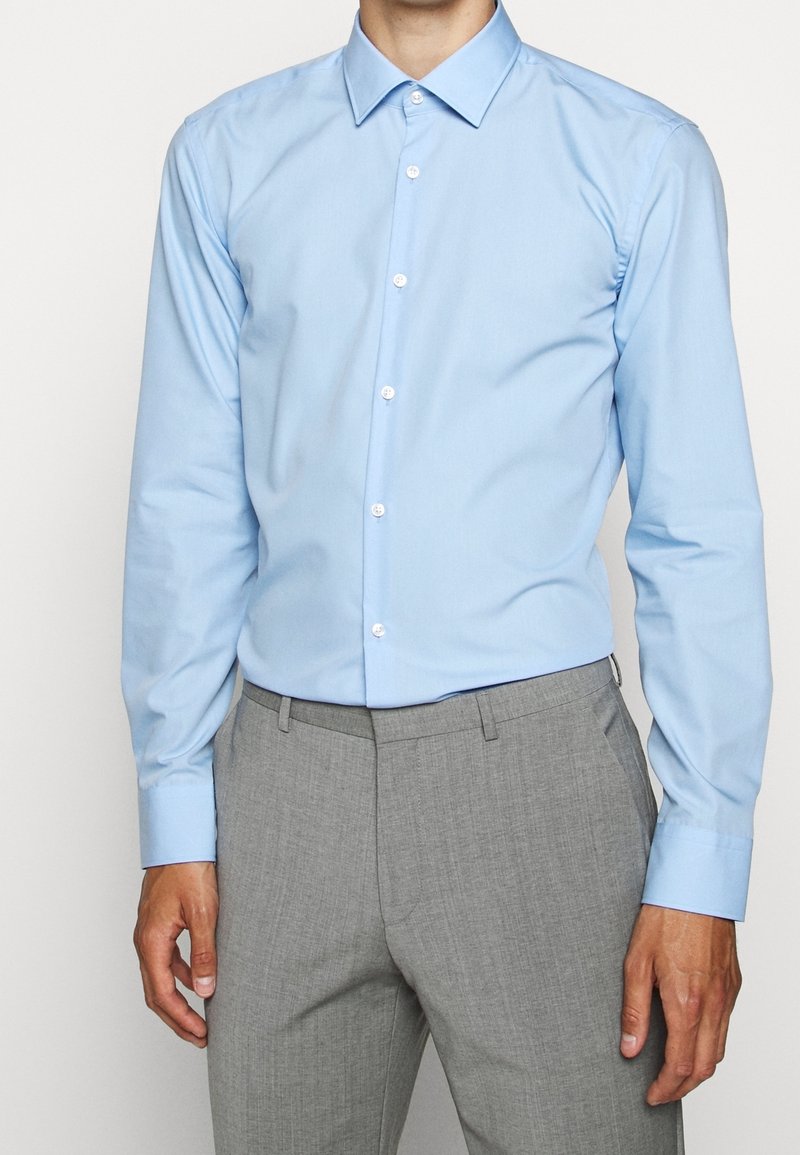Lichtblauw overhemd met lange mouwen, voorzien van een puntige kraag en witte knopen. Draagt een grijze dresscode broek met textuur.