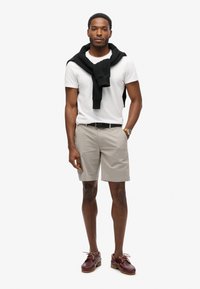 Lys grå shorts, hvid t-shirt, sort sweater bundet om halsen, brune sko, stående pose, minimalistisk baggrund.