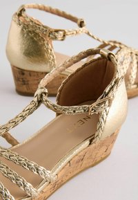 Goldene Keilsandalen mit geflochtenen Riemen, einer Korkplattform und einem glatten, metallischen Finish. Verstellbarer Schnallenverschluss enthalten.