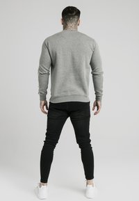 Mężczyzna z tatuażami na szyi i dłoniach, ubrany w szary sweter, czarne dżinsy i białe trampki, stojący tyłem na jednolitym tle.