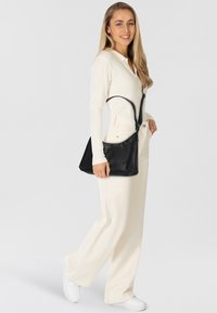 Borsa a tracolla in pelle nera con una texture liscia, forma triangolare e tracolla regolabile, indossata sopra una maglietta bianca a coste e pantaloni chiari.