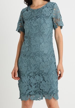 Kvinde iført en kortærmet, knælængde, teal blonder kjole med blomsterprint og rund hals mod en ensfarvet baggrund.