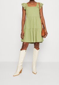 Groen losvallend zomerjurkje met laagjes en kanten accenten op de schouders, gecombineerd met witte kniehoge puntige laarzen en een kleine bruine crossbodytas.