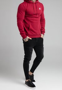 Rode hoodie met een voorkPocket en trekkoorden, gecombineerd met gescheurde zwarte skinny jeans en zwarte sneakers met witte zolen.
