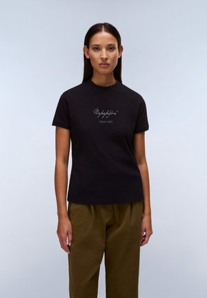 Frau mit glattem dunklem Haar, die ein schwarzes Napapijri-T-Shirt und olivgrüne Hosen trägt, vor einem hellgrauen Hintergrund stehend.