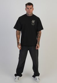 Juodas trumpas rankovė t-shirt su logotipu ant krūtinės, derinamas su tamsiai mėlynomis džinsinėmis kelnėmis ir baltomis sportbačiais. Modelis turi tatuiruotes ir neutralų požiūrį.