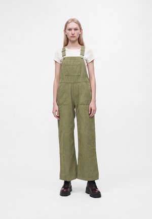 Donna con lunghi capelli biondi indossa una salopette di velluto verde, una camicia bianca a maniche corte e stivali neri con lacci, in piedi davanti a uno sfondo bianco semplice.