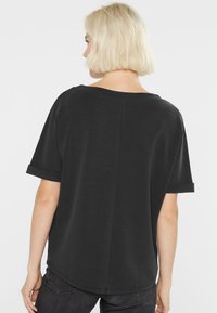 Person with short blonde hair wearing a loose black short-sleeve top and dark jeans, facing away against a plain white background.