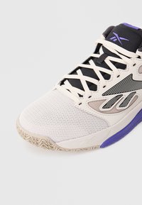 Biela a fialová športová obuv s priedušnou špičkou, čiernym jazykom, bielymi šnúrkami a čierno-bielym pruhovaným logom na boku.