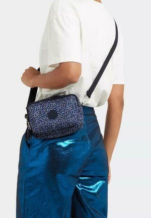 Persona che indossa una camicia bianca e pantaloni blu lucidi, con una piccola borsa a tracolla caratterizzata da una tracolla nera e un motivo blu a pois.