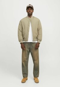 Veste bomber vert olive clair avec un design matelassé, t-shirt blanc en dessous, jeans bleus et bottes de travail beiges, accompagnée d'une casquette marron.
