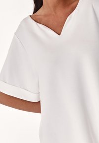 Weißes V-Ausschnitt-Top mit kurzen aufgerollten Ärmeln, aus glattem Stoff gefertigt und in einem sauberen, minimalistischen Design mit weicher Textur.