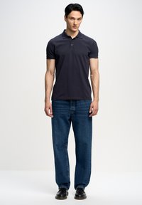 Tamsiai mėlynas polo marškinėliai su apykakle ir mygtukų juosta, derinami su mėlynomis tiesiomis džinsinėmis kelnėmis ir juodais batais. Lygaus audinio, standartinio kirpimo.