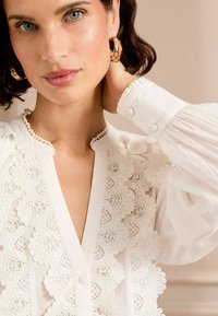 Blusa bianca con un dettagliato bordo in pizzo lungo il colletto e la patta, con maniche a sbuffo e polsini con bottoni. Tessuto di cotone morbido.